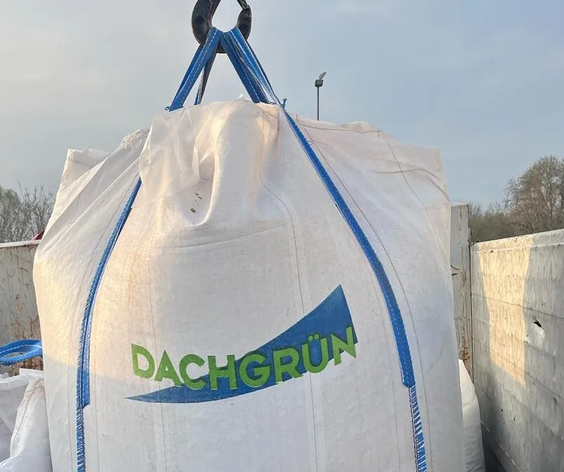 BigBag mit DACHGRÜN-Schriftzug in Grün auf blauem Keil hängt am Kranhaken